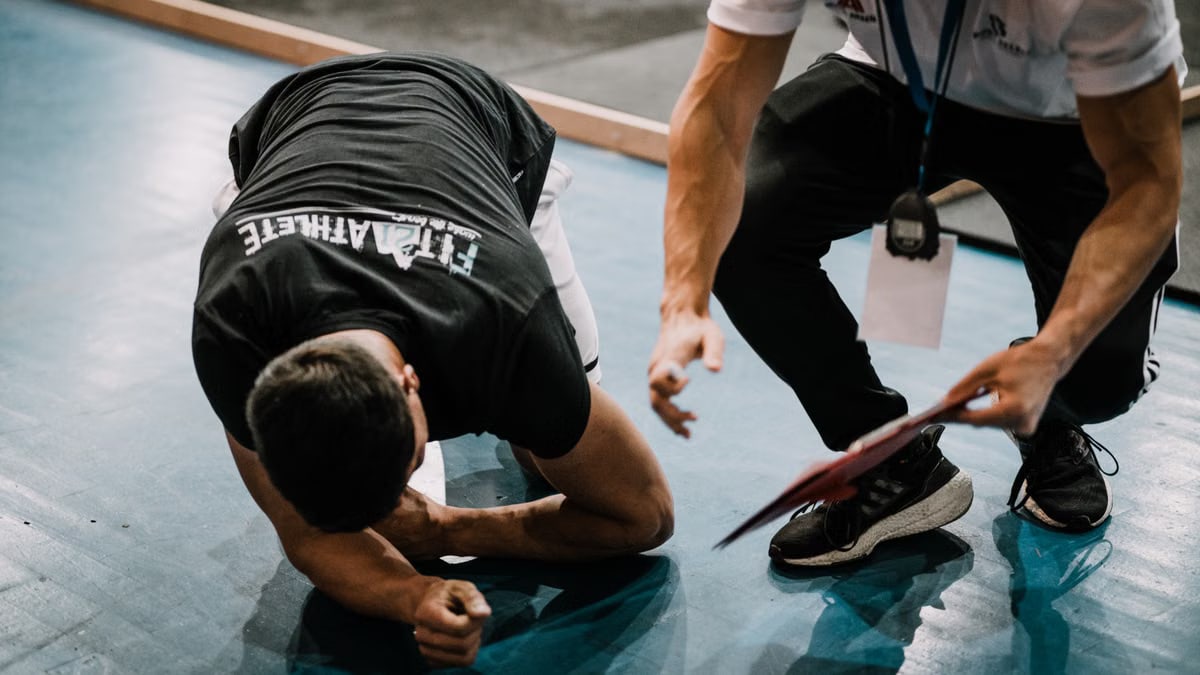 An athlete being assisted by a coach during an indoor training session on a blue floor.